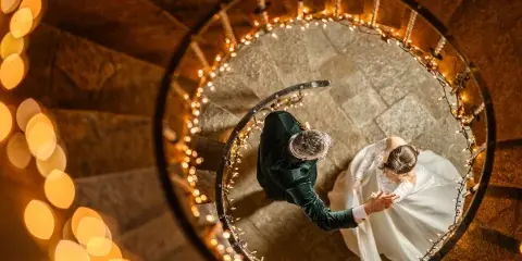 A bride and groom holding hands on a spiral staircase decorated with string lights, viewed from above.