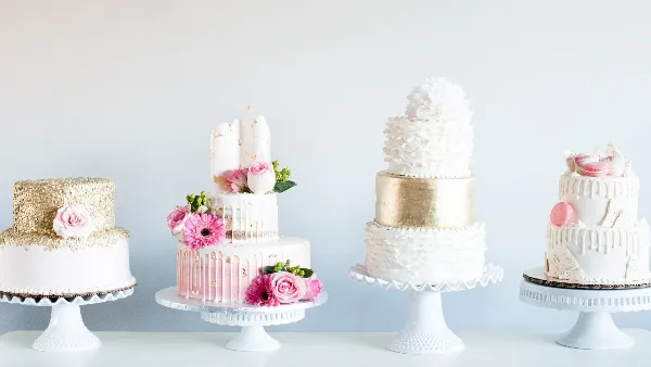 A display of four different multi-tiered wedding cakes featuring white and gold designs, fresh flowers, and decorative toppings.