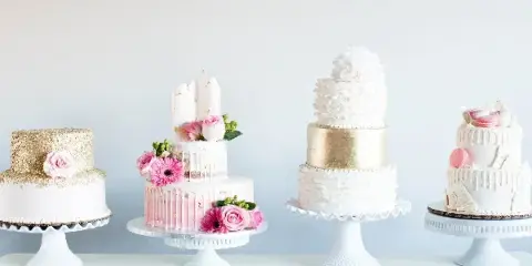 A display of four different multi-tiered wedding cakes featuring white and gold designs, fresh flowers, and decorative toppings.