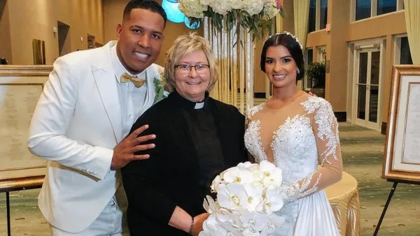 A wedding officiant in clerical attire poses with a bride and groom in a hotel ballroom.