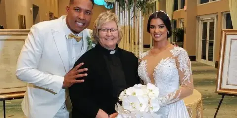 A wedding officiant in clerical attire poses with a bride and groom in a hotel ballroom.