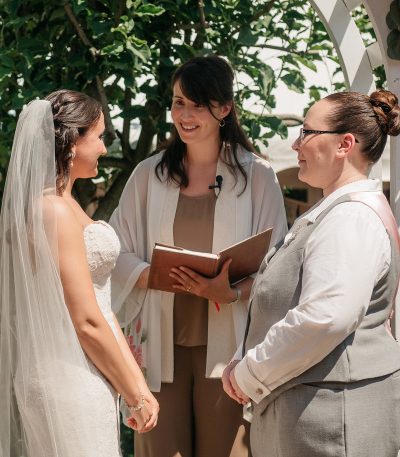 A wedding officiant stands between two brides during an outdoor ceremony, holding a book and smiling.