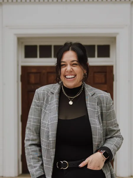 Portrait of a smiling woman in a plaid blazer standing in front of a white door.