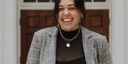 Portrait of a smiling woman in a plaid blazer standing in front of a white door.
