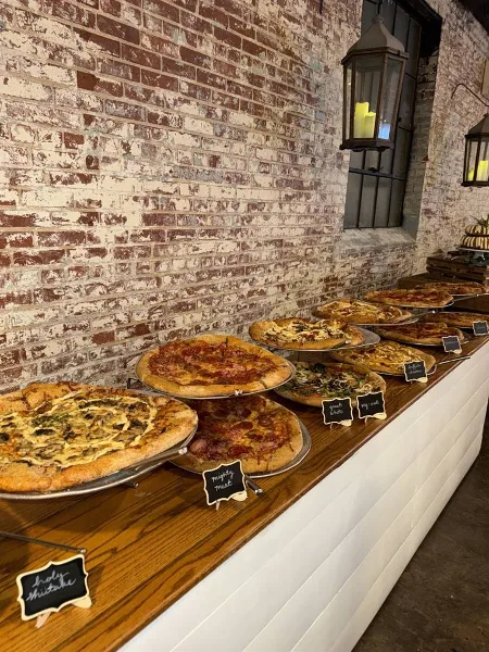 A row of stone-baked pizzas with handwritten name cards displayed on a wooden table against a brick wall.