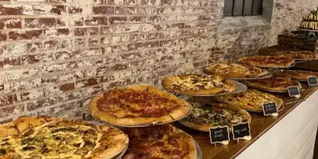 A row of stone-baked pizzas with handwritten name cards displayed on a wooden table against a brick wall.