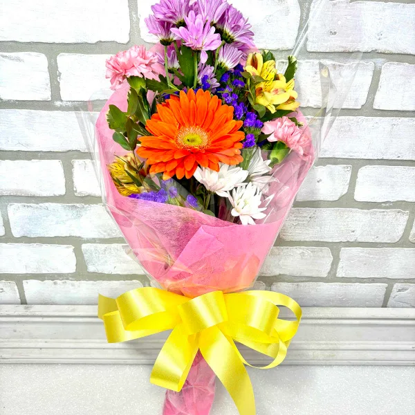A colorful mixed flower bouquet featuring a large orange gerbera daisy, wrapped in pink paper and tied with a yellow ribbon.
