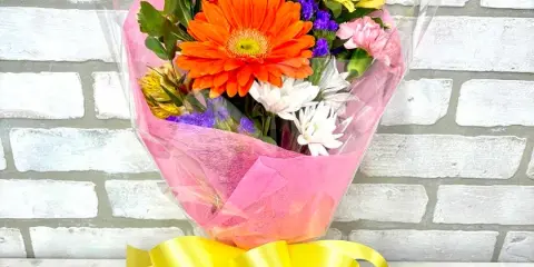 A colorful mixed flower bouquet featuring a large orange gerbera daisy, wrapped in pink paper and tied with a yellow ribbon.