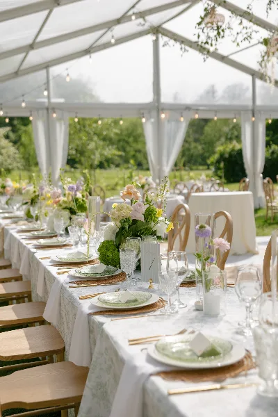 A long wedding reception table set with white linens, green plates, and floral centerpieces inside a white tent.