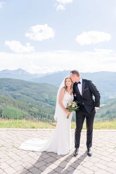 A bride and groom standing on a paved overlook with a mountain range in the background.