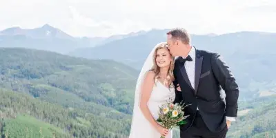 A bride and groom standing on a paved overlook with a mountain range in the background.