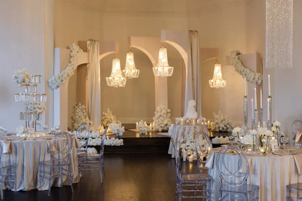 A luxury wedding venue interior featuring a custom archway installation, crystal chandeliers, white floral arrangements, and clear acrylic chairs.