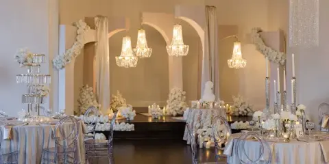 A luxury wedding venue interior featuring a custom archway installation, crystal chandeliers, white floral arrangements, and clear acrylic chairs.