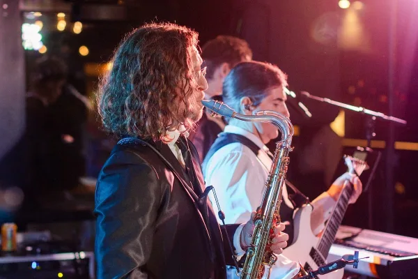 A live band performing on stage, featuring a saxophone player in the foreground and a guitarist in the background.