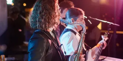 A live band performing on stage, featuring a saxophone player in the foreground and a guitarist in the background.