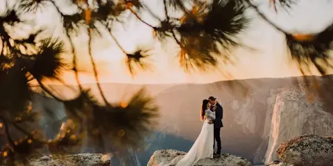 A bride and groom embracing on a rocky outcrop in Yosemite National Park at sunset, framed by pine branches.