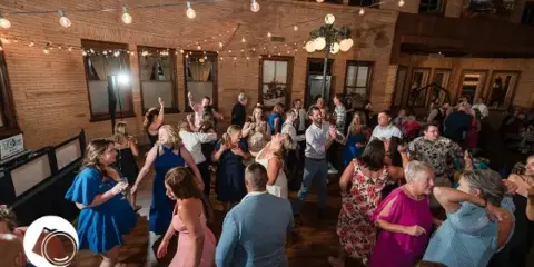Guests dancing at a wedding reception in a venue with string lights and brick walls.