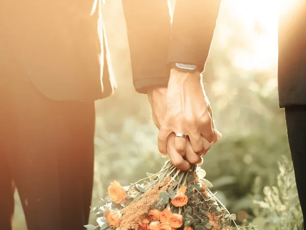 A close-up of two people holding hands, one wearing a wedding ring, with a bouquet of orange roses and greenery held below.