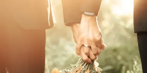 A close-up of two people holding hands, one wearing a wedding ring, with a bouquet of orange roses and greenery held below.