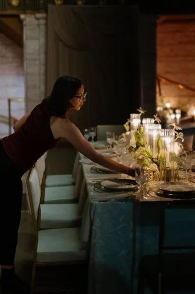 A wedding planner arranging a reception table with linens, candles, and floral centerpieces.