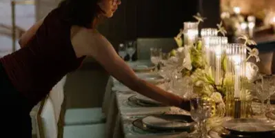 A wedding planner arranging a reception table with linens, candles, and floral centerpieces.