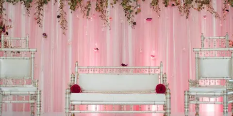 Wedding lounge setup with pink draping, floral garland, and white upholstered seating.