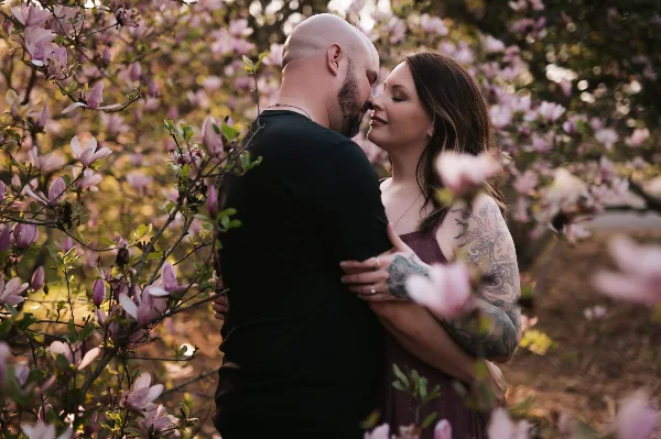 A professional photograph of a couple embracing and touching foreheads amidst blooming pink magnolia flowers.