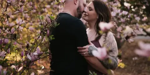 A professional photograph of a couple embracing and touching foreheads amidst blooming pink magnolia flowers.