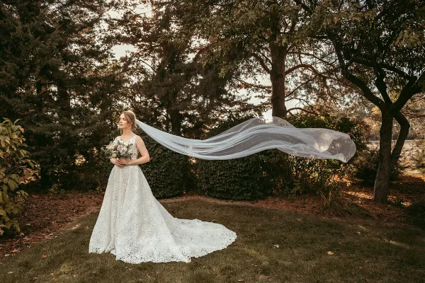A bride in a white lace wedding dress stands in a wooded area holding a bouquet, with her long veil blowing in the wind.