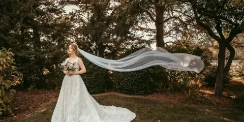 A bride in a white lace wedding dress stands in a wooded area holding a bouquet, with her long veil blowing in the wind.