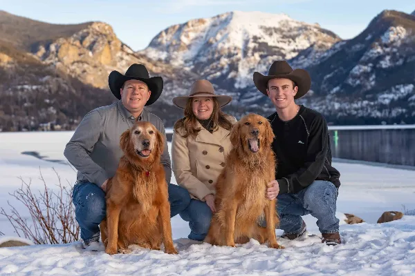 A family of three wearing cowboy hats poses with two golden retrievers in the snow with mountains in the background.