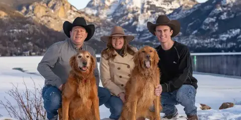 A family of three wearing cowboy hats poses with two golden retrievers in the snow with mountains in the background.