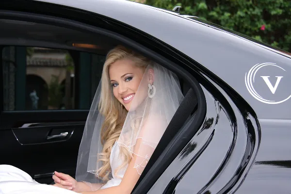 A smiling bride in a white wedding dress and veil sitting in the back seat of a black luxury sedan with a Vitesse logo on the door.