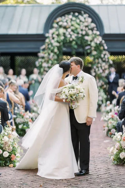 A bride and groom share a kiss at a wedding ceremony featuring a large floral arch and aisle arrangements.
