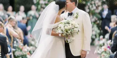 A bride and groom share a kiss at a wedding ceremony featuring a large floral arch and aisle arrangements.