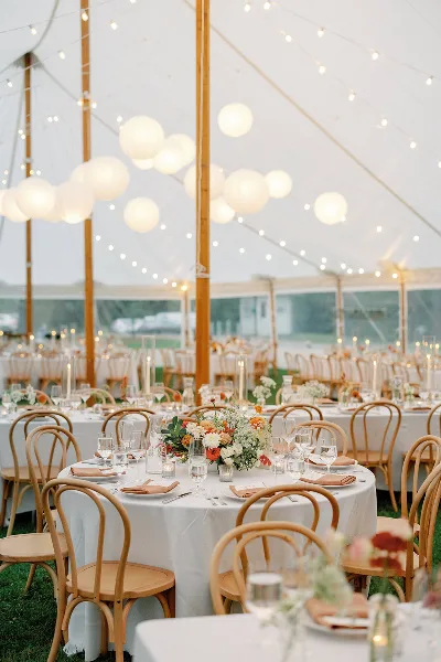 A styled wedding reception inside a white tent featuring round tables with white linens, wooden chairs, floral centerpieces, and hanging paper lanterns.