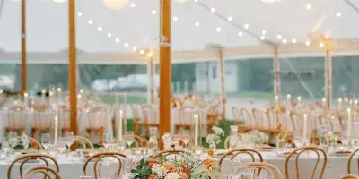 A styled wedding reception inside a white tent featuring round tables with white linens, wooden chairs, floral centerpieces, and hanging paper lanterns.
