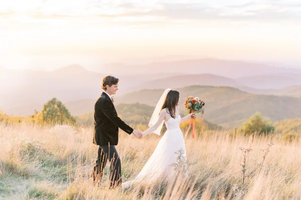 A bride and groom holding hands and walking through a golden grassy field with mountains in the background.