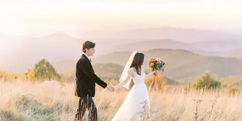 A bride and groom holding hands and walking through a golden grassy field with mountains in the background.