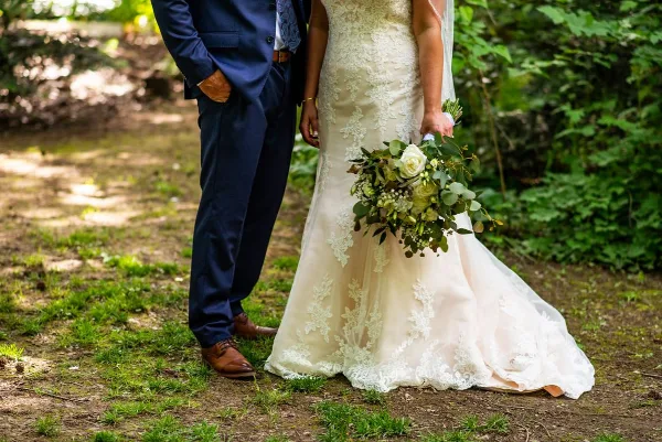 A bride in a lace wedding dress holding a bouquet and a groom in a navy suit standing outdoors in a wooded area.
