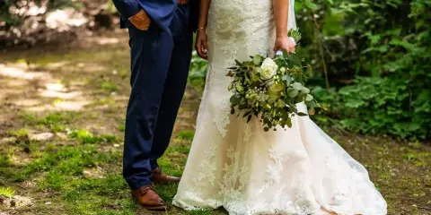 A bride in a lace wedding dress holding a bouquet and a groom in a navy suit standing outdoors in a wooded area.