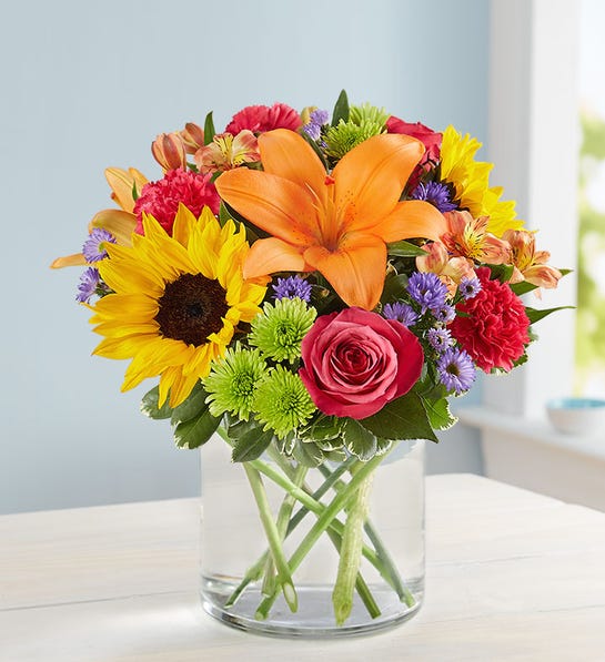 A colorful mixed flower bouquet featuring a sunflower, orange lily, and pink rose in a clear glass vase.