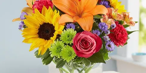 A colorful mixed flower bouquet featuring a sunflower, orange lily, and pink rose in a clear glass vase.