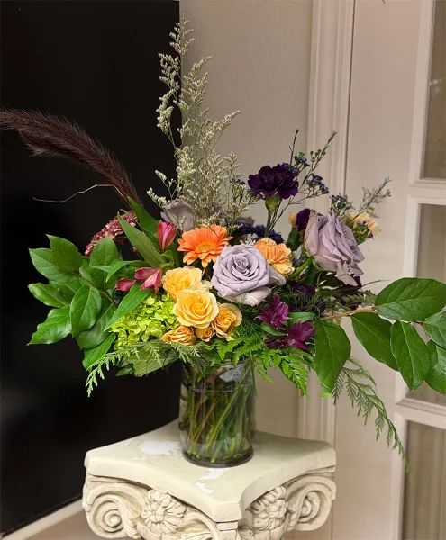 A colorful floral arrangement with yellow and purple roses, orange daisies, and greenery in a clear glass vase on a decorative pedestal.