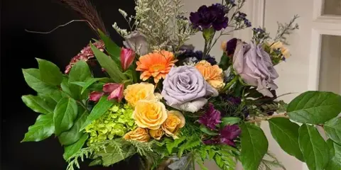 A colorful floral arrangement with yellow and purple roses, orange daisies, and greenery in a clear glass vase on a decorative pedestal.