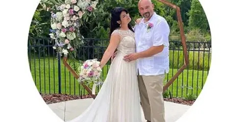 A bride and groom posing in front of a floral wedding archway with a bouquet.