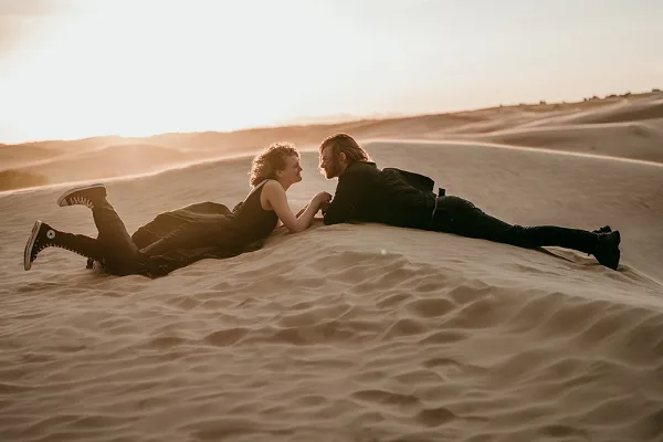 A couple lying face-to-face on a sand dune at golden hour, photographed in a professional portrait style.