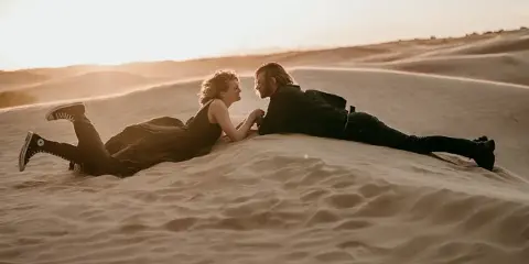 A couple lying face-to-face on a sand dune at golden hour, photographed in a professional portrait style.