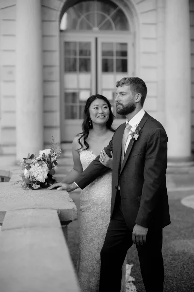 A black and white photograph of a bride and groom standing together outdoors in front of a stone building.