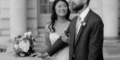 A black and white photograph of a bride and groom standing together outdoors in front of a stone building.